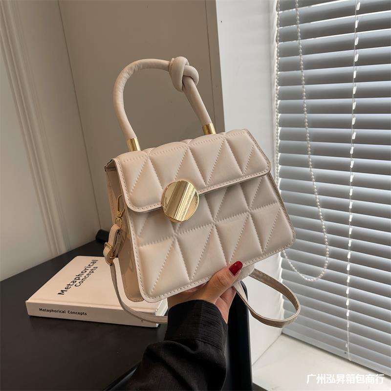 Beige quilted handbag with gold accents held by a person, with a book and window blinds in the background.