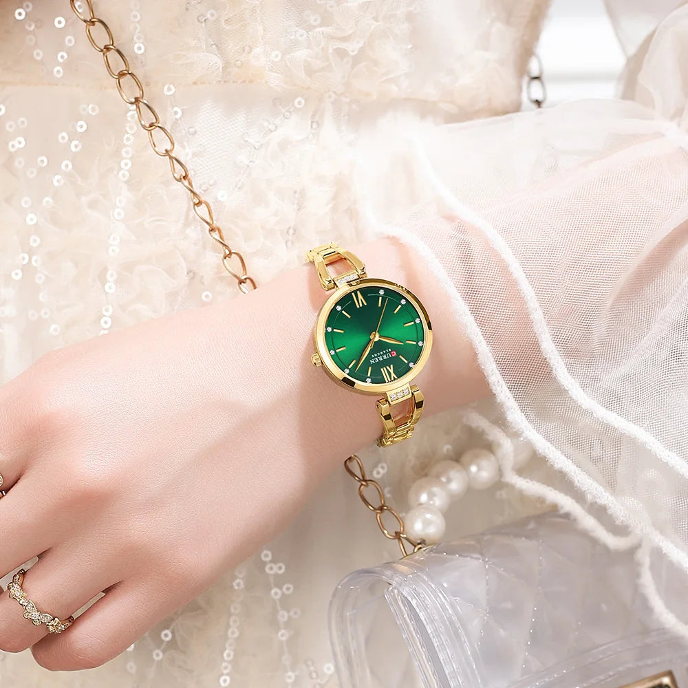 Hand wearing a gold watch with a green face, against a light background.