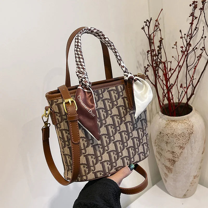 Designer handbag with monogram pattern held by a person, next to a decorative vase with branches.